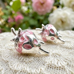 May include: A pair of silver-toned earrings with a unique design. Each earring features a pink enamel pattern with a glossy finish. The earrings have a three-dimensional shape with curved lines and a smooth, polished surface.