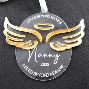 May include: Clear acrylic ornament featuring gold-coloured angel wings and a halo. The ornament is inscribed with the word "Nanny" and the year "2025" in white. The phrases "LOVED BEYOND WORDS" and "MISSED BEYOND MEASURE" are also printed on the ornament.