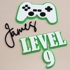 May include: Gaming-themed cake toppers in white and green. Features a controller, the name "James" in black script, and the words "LEVEL 9" in white letters with a green border. Ideal for a birthday cake.