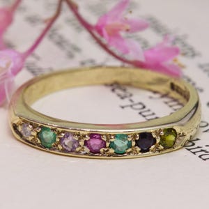 May include: A gold band ring featuring a row of six small, round, multi-colored gemstones in shades of green, purple, pink, and dark blue. The ring is positioned in front of a small sprig of light pink flowers with thin, brown stems.
