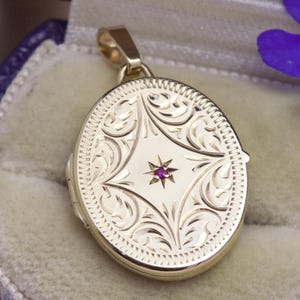 May include: An oval gold locket pendant featuring an intricate engraved scrollwork pattern and a central star-shaped setting containing a small, vibrant pink gemstone. The locket has a polished finish and a small bail at the top for attachment.