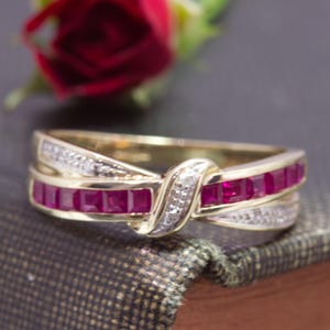 May include: A gold ring featuring a crossover design with a row of square-cut red rubies and two rows of small, clear accent stones. The band has a polished finish and the stones are set in a bypass style that creates a central knot-like detail.