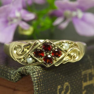May include: A gold ring with a central diamond-shaped setting holding four red gemstones. The ring is embellished with small, round pearls and ornate, swirling gold filigree. The ring is set against a backdrop of purple flowers.