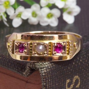 以下が含まれることがあります： 3つの赤い宝石と中央に1つの真珠が付いた金の指輪。指輪は茶色の本の上に置かれています。