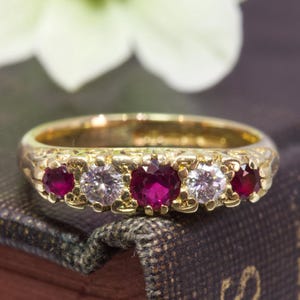 以下が含まれることがあります： 繊細な花の模様に、3つのルビーと2つのダイヤモンドがセットされたゴールドのバンドリングです。
