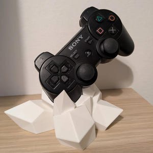 Puede incluir: Un mando de PlayStation 3 negro se muestra en un soporte geométrico blanco. El mando tiene la palabra "SONY" impresa en él.