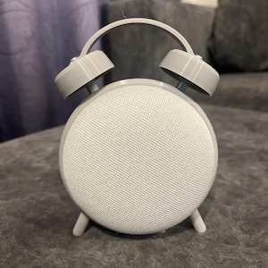 May include: A gray, retro-style alarm clock with a round, speaker-like face and two bell-shaped alarms on top. The clock has a curved handle and three small legs. The face is covered in a textured, light gray fabric.