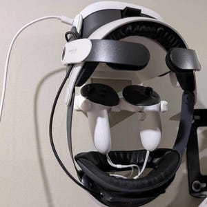 Puede incluir: Un auricular de realidad virtual blanco con correas negras y controladores está montado en una pared. El auricular tiene un acolchado de espuma negra y un cable blanco conectado a él.
