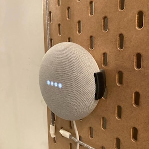 Puede incluir: Un altavoz inteligente Google Home Mini gris montado en una pared de tablero de clavijas con un cable blanco enchufado. El altavoz tiene una cubierta de tela gris claro y una fila de cuatro luces blancas en la parte delantera.