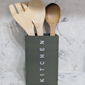 Könnte beinhalten: Ein dunkelgrüner Utensilienhalter mit dem Wort "KITCHEN" in weiß auf der Vorderseite. Drei Holzlöffel befinden sich im Halter.