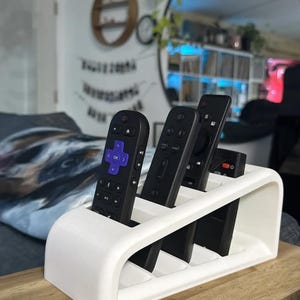 May include: White plastic remote control organizer with five slots for remotes. The organizer is angled and has a curved front. Five black remotes are in the slots.