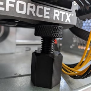 May include: Close-up of a black computer component support screw. The screw is supporting a GeForce RTX graphics card. Yellow and black cables are visible in the background. The screw has a textured top for easy adjustment.