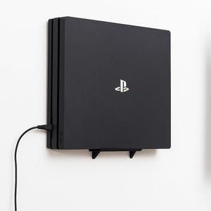 May include: A black PlayStation 4 console mounted on a white wall. The console is rectangular with a gold and white logo. A black power cable is plugged into the console and hangs down. The console is supported by a black bracket.