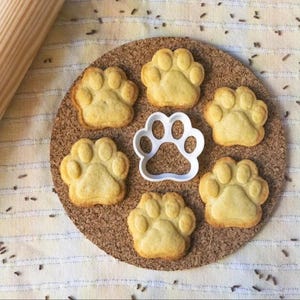 May include: Six light brown paw print cookies on a cork coaster with a white paw print cookie cutter in the center.