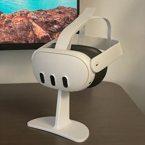 Puede incluir: Soporte de auriculares VR blanco con una base blanca y una correa blanca. Los auriculares están apoyados en el soporte.