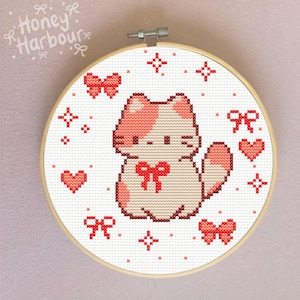 Peut inclure: Un tambour de broderie au point de croix avec un motif de chat. Le chat est beige clair et rose, avec un nœud rouge et entouré de nœuds, de cœurs et d'étoiles rouges. Le texte "Honey Harbour" est dans le coin supérieur gauche.