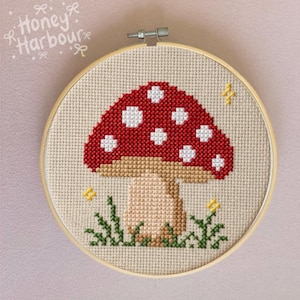 Puede incluir: Un bordado de punto de cruz en un aro de madera presenta una seta roja con lunares blancos. La seta se encuentra sobre un fondo beige con hierba verde y acentos de estrellas amarillas. El texto "Honey Harbour" está en la esquina superior izquierda.