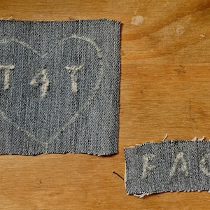May include: Two denim patches with white lettering. The first patch is a heart shape with the word "TAT" inside. The second patch is rectangular with the word "FAG" inside.