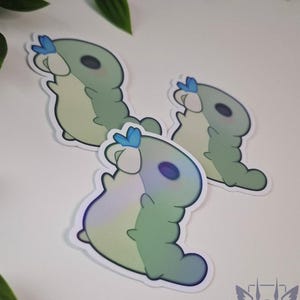 Puede incluir: Tres pegatinas con un diseño de oruga de dibujos animados. Las orugas son de color verde claro y blanco con detalles en azul. Las pegatinas tienen un acabado brillante y están dispuestas sobre una superficie blanca.