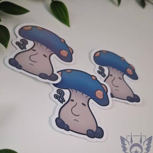Puede incluir: Tres pegatinas de hongos caprichosos con tapas azules y tallos de color marrón claro están dispuestas sobre una superficie blanca. Las tapas de los hongos tienen manchas de color marrón claro. Cada hongo tiene una cara con una expresión neutra. Las pegatinas son de Lynniia.