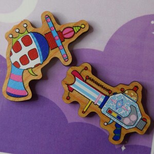 May include: Two wooden laser gun pins with colourful designs. One gun has a red and white grid pattern, while the other features a blue and white floral design. The pins are on a purple background.