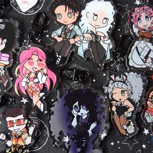 May include: A collection of cartoon character keychains with various designs. The keychains feature characters with pink, black, and white hair, wearing outfits in various colors. The background is black with white stars and constellations.