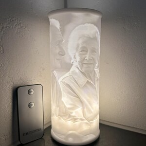 May include: A white cylindrical lamp with a portrait of an elderly person is illuminated from within. A remote control with "ON" and "OFF" buttons and "REMOTE CONTROL" text is next to the lamp. The lamp is on a dark surface.