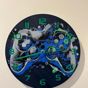 May include: A blue and black circular wall clock with a video game controller design. The clock face has green numbers and a green second hand.