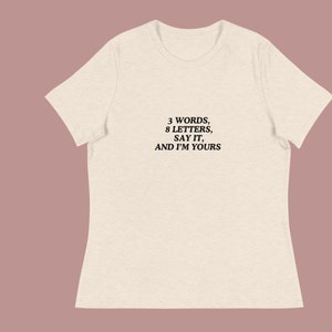 Może przedstawiać: Beżowa koszulka z nadrukiem "3 WORDS, 8 LETTERS, SAY IT, AND I'M YOURS" w kolorze czarnym.