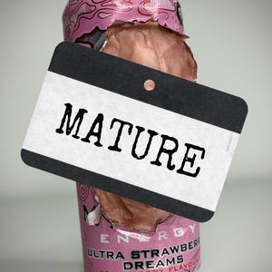 Puede incluir: Una lata de bebida energética rosa aplastada con el texto "ULTRA STRAWBERRY DREAMS" y "ZERO SUGAR". Una etiqueta negra con la palabra "MATURE" está delante de la lata.