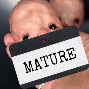 Könnte beinhalten: Zwei blass, fleischfarbene, geformte Köpfe mit schwarzen Augen sind teilweise sichtbar. Ein schwarz-weißes Etikett mit dem Wort "MATURE" in fetten, schwarzen Buchstaben wird vor den Köpfen gehalten. Das Etikett hat einen roten Punkt.