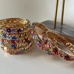 Może przedstawiać: Złote bransoletki z misternymi wzorami kwiatowymi. Bransoletki zdobią kolorowe emaliowane kwiaty w odcieniach różu, błękitu, fioletu i bieli. Niektóre są ułożone w stos, inne wyeksponowane osobno, ukazując szczegółowe rzemiosło.