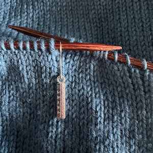 May include: A close-up of a knitting project with a silver ruler charm hanging from the needles. The wooden needles are dark, and the yarn is a textured teal colour. The ruler charm displays measurements in centimetres.