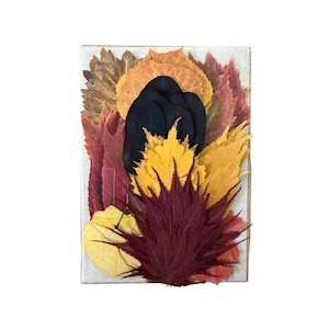 May include: A rectangular canvas featuring a layered collage of pressed autumn leaves in shades of deep burgundy, vibrant yellow, burnt orange, and dark brown, with a prominent black leaf centered among the colorful foliage arrangement.