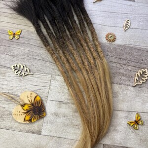 Peut inclure: Un ensemble de dreadlocks ombrés noirs et blonds. Les dreadlocks passent du noir aux racines au blond aux pointes. Des découpes en bois de papillons et de feuilles sont dispersées autour des dreadlocks, ainsi qu'un cœur en bois avec un motif de tournesol.