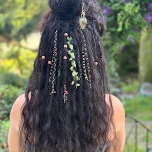 May include: Dark, wavy hair styled in a bun, adorned with green leaf accents, wooden beads, and gold-colored spiral hair wraps. A gold-colored charm hangs from the bun. The hair is styled with a natural, bohemian aesthetic.