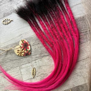 May include: A bundle of vibrant, handmade dreadlocks. The dreadlocks transition from black at the roots to a bright pink hue. The image is set against a light gray wooden surface, with decorative wooden leaf and heart accents.