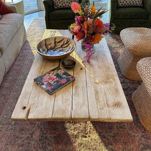 May include: A wooden coffee table with a natural finish, featuring a bowl with a black and white design, a small bowl, and a book titled "Color Me Floral". The table is surrounded by a colorful rug, a green armchair with a patterned pillow, and a woven stool.