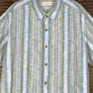May include: A short-sleeved button-down shirt with vertical stripes in white, light blue, and green. The shirt has a chest pocket and wooden-colored buttons. The brand name "Tommy Bahama" is visible on the collar.