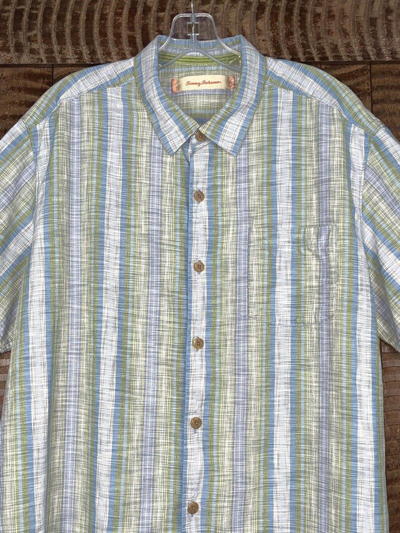 May include: A short-sleeved button-down shirt with vertical stripes in white, light blue, and green. The shirt has a chest pocket and wooden-colored buttons. The brand name "Tommy Bahama" is visible on the collar.
