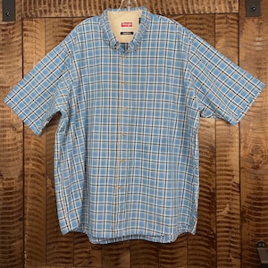 Pode incluir: Uma camisa de manga curta com botões em um padrão xadrez azul e branco. A camisa tem uma gola abotoada e um forro de gola bege. A marca "Wrangler" é visível na etiqueta.