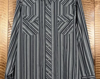 May include: A long-sleeved, button-down shirt with a black and white striped pattern. The shirt features two chest pockets and a collar. The Wrangler brand label is visible at the collar. The shirt is hanging on a hanger.