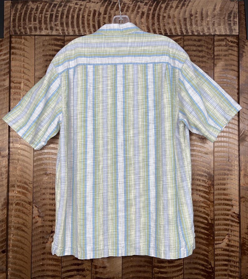 May include: Short-sleeved button-down shirt with a vertical striped pattern in white, light blue, and green. The shirt is hanging on a hanger against a wooden background. The back of the shirt is visible.
