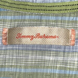 May include: Close-up of a Tommy Bahama shirt label. The label is rectangular with the brand name embroidered in red. The shirt has a blue and green plaid pattern with a green collar.
