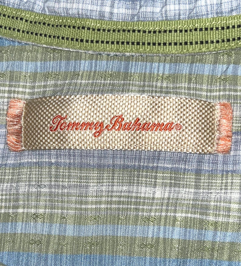 May include: Close-up of a Tommy Bahama shirt label. The label is rectangular with the brand name embroidered in red. The shirt has a blue and green plaid pattern with a green collar.