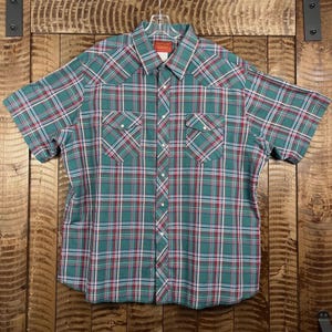 Puede incluir: Camisa de manga corta a cuadros verdes, rojos y blancos. Cuenta con dos bolsillos en el pecho y cierres de botones a presión de perlas. La camisa tiene un estilo occidental clásico.