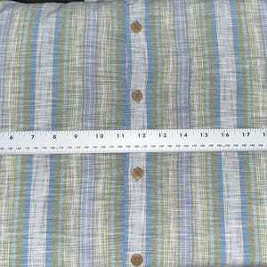 May include: A button-down shirt with a blue, green, and white striped pattern. The shirt is measured with a ruler, showing a width of approximately 24 inches. The shirt has wooden buttons.