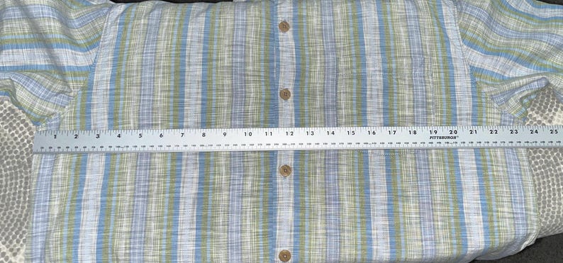 May include: A button-down shirt with a blue, green, and white striped pattern. The shirt is measured with a ruler, showing a width of approximately 24 inches. The shirt has wooden buttons.