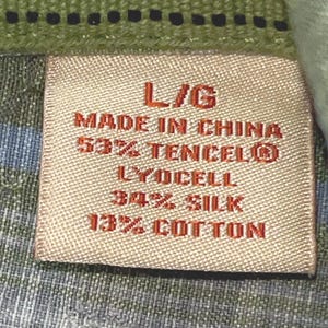 May include: Close-up of a clothing label with the text "L/G MADE IN CHINA 53% TENCEL&reg; LYOCELL 34% SILK 13% COTTON". The label is rectangular and beige, set against a green and blue patterned fabric.