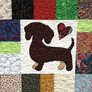 Pu&ograve; includere: Una trapunta patchwork con un'applicazione di bassotto marrone e un cuore. La trapunta &egrave; composta da vari quadrati di tessuto nei toni del rosso, verde, blu e bianco, con disegni floreali e fantasia. Il cane &egrave; lungo circa 25 cm.